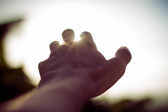 A hand reaching toward a bright light source, with sun rays filtering through the fingers, creating a sense of aspiration or longing.