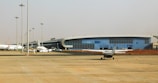 A modern airport terminal with a sleek, curved architectural design and large glass windows. Several airplanes, including a small aircraft, are parked on the tarmac. The open area in the foreground shows a few safety cones and light poles, with a clear sky in the background.