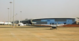 A modern airport terminal with a sleek, curved architectural design and large glass windows. Several airplanes, including a small aircraft, are parked on the tarmac. The open area in the foreground shows a few safety cones and light poles, with a clear sky in the background.