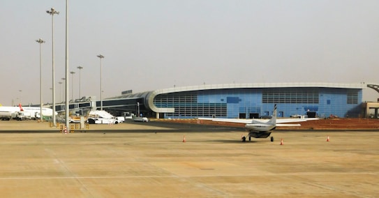 A modern airport terminal with a sleek, curved architectural design and large glass windows. Several airplanes, including a small aircraft, are parked on the tarmac. The open area in the foreground shows a few safety cones and light poles, with a clear sky in the background.