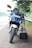 A blue and white motorcycle is parked on the side of the road next to a guardrail, with a black bag hanging from the handlebar under a tree. The license plate is visible and the setting is outdoors, with greenery in the background.