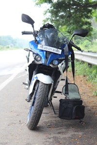 A blue and white motorcycle is parked on the side of the road next to a guardrail, with a black bag hanging from the handlebar under a tree. The license plate is visible and the setting is outdoors, with greenery in the background.