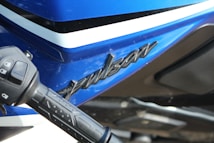 Close-up view of a motorcycle with a blue, black, and white color scheme. The word 'Pulsar' is prominently displayed on the side panel. A handlebar grip is visible in the foreground, featuring various control buttons.