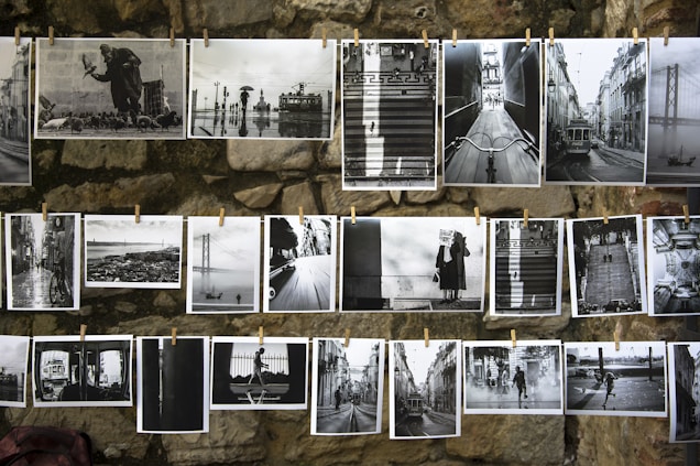A collection of black and white photographs is displayed on a stone wall, each pinned by clothespins. The images showcase various cityscapes, urban scenes, and people, including trams, bridges, and figures with umbrellas. The overall presentation emphasizes contrast and composition.