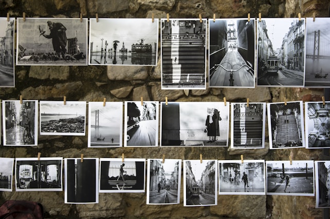 A collection of black and white photographs is displayed on a stone wall, each pinned by clothespins. The images showcase various cityscapes, urban scenes, and people, including trams, bridges, and figures with umbrellas. The overall presentation emphasizes contrast and composition.