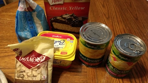 A variety of grocery items are displayed on a wooden table. There is a box of yellow cake mix, a bag of shredded coconut, a tub of butter spread, a bag of sliced almonds, and two cans of fruit, one labeled crushed pineapple and the other fruit cocktail. The items appear to be ingredients for baking or making a dessert.