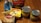 A variety of grocery items are displayed on a wooden table. There is a box of yellow cake mix, a bag of shredded coconut, a tub of butter spread, a bag of sliced almonds, and two cans of fruit, one labeled crushed pineapple and the other fruit cocktail. The items appear to be ingredients for baking or making a dessert.
