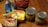 A variety of grocery items are displayed on a wooden table. There is a box of yellow cake mix, a bag of shredded coconut, a tub of butter spread, a bag of sliced almonds, and two cans of fruit, one labeled crushed pineapple and the other fruit cocktail. The items appear to be ingredients for baking or making a dessert.