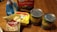 A variety of grocery items are displayed on a wooden table. There is a box of yellow cake mix, a bag of shredded coconut, a tub of butter spread, a bag of sliced almonds, and two cans of fruit, one labeled crushed pineapple and the other fruit cocktail. The items appear to be ingredients for baking or making a dessert.