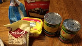 A variety of grocery items are displayed on a wooden table. There is a box of yellow cake mix, a bag of shredded coconut, a tub of butter spread, a bag of sliced almonds, and two cans of fruit, one labeled crushed pineapple and the other fruit cocktail. The items appear to be ingredients for baking or making a dessert.