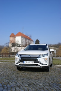A white Mitsubishi SUV is parked on a cobblestone driveway with a historic building featuring a red roof in the background. The sky is clear and blue, and trees without leaves are visible, suggesting a winter or early spring setting.