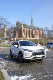 A white Mitsubishi Eclipse Cross is parked on a cobblestone pavement in front of a historic church. The church features a tall spire and is surrounded by bare trees, indicating a winter or early spring setting. The sky is clear and blue, creating a bright atmosphere.