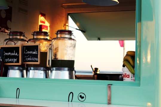 A food truck counter with glass jars labeled 'Limonada Tradicional' and 'Limonada de Orange' on a metal stand. There is a container of Nutella with bananas placed on top of it. The counter is teal with a simple design and appears to be set against a backdrop of an open serving window.