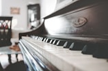 A close-up view of a piano keyboard in a softly lit room, focusing on the keys with a blurred background including furniture like chairs and a cabinet.