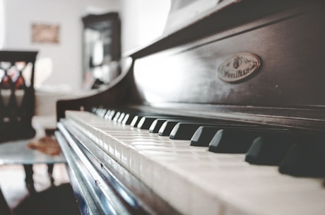 A close-up view of a piano keyboard in a softly lit room, focusing on the keys with a blurred background including furniture like chairs and a cabinet.
