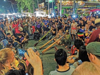 A vibrant street scene with a large diverse crowd gathered around several musicians playing long, wind instruments. The audience appears engaged and many are taking photos or videos with their smartphones. The setting is likely a night market or festival with bright lights and food stalls in the background.