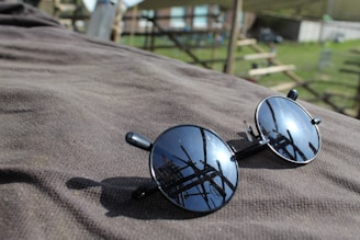 A pair of round sunglasses with dark lenses is placed on a textured surface, reflecting the surrounding structures. The background is partially blurred, showing elements of an outdoor setting with some wooden frames and greenery.