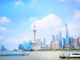 A vibrant city skyline featuring modern skyscrapers along a waterfront. The iconic Oriental Pearl Tower stands prominently among various high-rise buildings under a clear blue sky. Boats are seen moving along the river in the foreground, adding life to the urban scene.