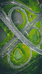 An aerial view of a complex highway interchange with multiple curved roads and loops. There are patches of greenery and small bodies of water surrounding the roads, creating an integrated natural landscape within the urban environment.