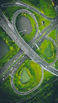 An aerial view of a complex highway interchange with multiple curved roads and loops. There are patches of greenery and small bodies of water surrounding the roads, creating an integrated natural landscape within the urban environment.