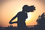 A silhouette of a person dancing with flowing hair in front of a bright, setting sun. The background includes trees and buildings under a clear sky, creating a serene and artistic atmosphere.