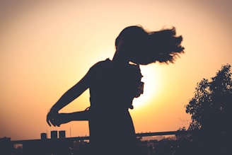 A silhouette of a person dancing with flowing hair in front of a bright, setting sun. The background includes trees and buildings under a clear sky, creating a serene and artistic atmosphere.