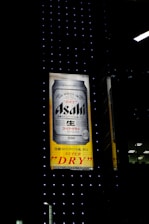 An illuminated billboard displays an advertisement for Asahi Super Dry beer, featuring a large image of a beer can against a background of black with small dotted lights. Japanese text is visible alongside the brand name.