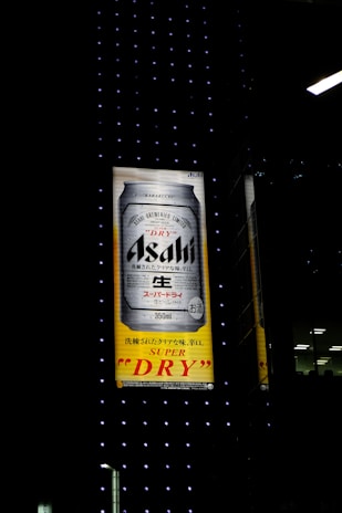 An illuminated billboard displays an advertisement for Asahi Super Dry beer, featuring a large image of a beer can against a background of black with small dotted lights. Japanese text is visible alongside the brand name.