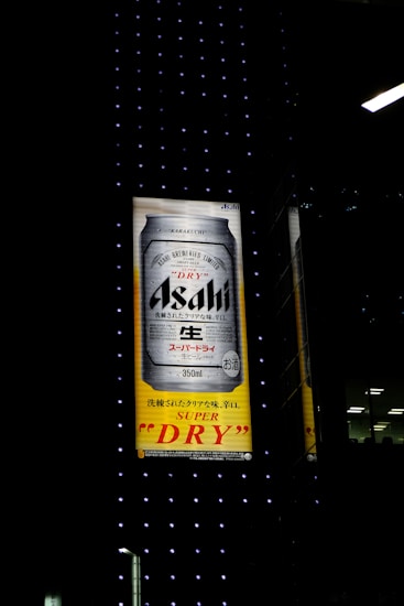 An illuminated billboard displays an advertisement for Asahi Super Dry beer, featuring a large image of a beer can against a background of black with small dotted lights. Japanese text is visible alongside the brand name.