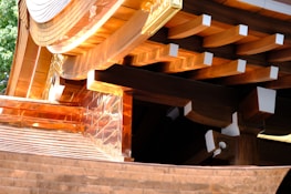 A detailed view of wooden architectural elements featuring smooth, curved surfaces and precisely cut beams. Copper or bronze metalwork adds a reflective quality to the structure, with sunlight enhancing the warm tones. The craftsmanship highlights traditional construction techniques.