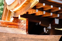 A detailed view of wooden architectural elements featuring smooth, curved surfaces and precisely cut beams. Copper or bronze metalwork adds a reflective quality to the structure, with sunlight enhancing the warm tones. The craftsmanship highlights traditional construction techniques.