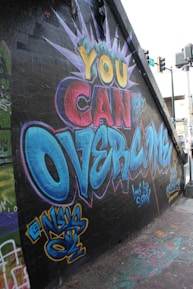 Vibrant graffiti on a wall features motivational text in large, colorful letters. The words 'You Can Overcome' are prominently displayed, designed with dynamic colors including yellow, red, and blue. The artistic style includes bold outlines and a burst design surrounding the text. Various tags and smaller pieces of graffiti art can be seen decorating the surrounding wall and ground area.