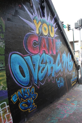 Vibrant graffiti on a wall features motivational text in large, colorful letters. The words 'You Can Overcome' are prominently displayed, designed with dynamic colors including yellow, red, and blue. The artistic style includes bold outlines and a burst design surrounding the text. Various tags and smaller pieces of graffiti art can be seen decorating the surrounding wall and ground area.