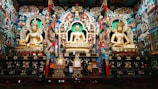 An ornate temple interior with three large golden statues of seated figures, richly decorated with intricate patterns and colorful designs. The walls are adorned with detailed murals and vibrant motifs, adding to the grandeur and spiritual ambiance. Lavish use of gold and embellishments highlights the cultural and religious significance of the space.