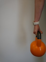 A person's hand is holding an orange kettlebell by its black handle. The person's wrist has a white smartwatch band. The background is plain and neutral.