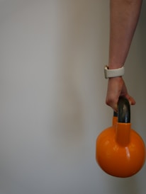 A person's hand is holding an orange kettlebell by its black handle. The person's wrist has a white smartwatch band. The background is plain and neutral.