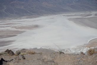 An expansive desert landscape featuring a vast salt flat surrounded by rocky hills. The foreground contains sparse vegetation and rough terrain, while the background reveals distant mountain ranges with intricate patterns etched into their slopes.