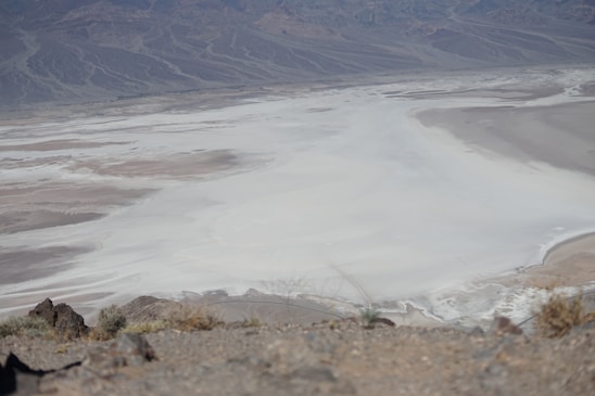 An expansive desert landscape featuring a vast salt flat surrounded by rocky hills. The foreground contains sparse vegetation and rough terrain, while the background reveals distant mountain ranges with intricate patterns etched into their slopes.