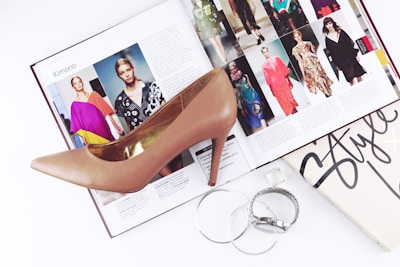 A fashion magazine is open to a page featuring various models wearing colorful outfits. A high-heeled shoe is placed on top of the magazine. Beside it, silver jewelry including a bracelet and two bangles are arranged, along with a book titled 'Style'.
