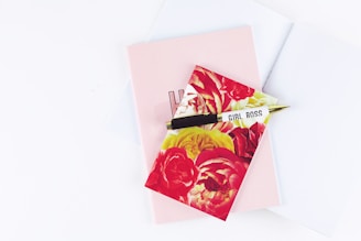 A floral-patterned notepad with large red and yellow flowers is placed on top of a plain pink notebook. A gold and black pen, inscribed with 'GIRL BOSS,' rests diagonally across them. The background is a soft white surface.