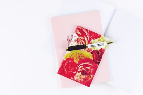 A floral-patterned notepad with large red and yellow flowers is placed on top of a plain pink notebook. A gold and black pen, inscribed with 'GIRL BOSS,' rests diagonally across them. The background is a soft white surface.