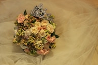 A bouquet of roses and other flowers in soft pink and cream hues is adorned with a decorative silver tiara. The background features layers of sheer, delicate fabric that resemble tulle, enhancing the romantic, elegant feel of the arrangement.