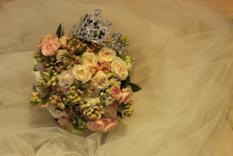 A bouquet of roses and other flowers in soft pink and cream hues is adorned with a decorative silver tiara. The background features layers of sheer, delicate fabric that resemble tulle, enhancing the romantic, elegant feel of the arrangement.