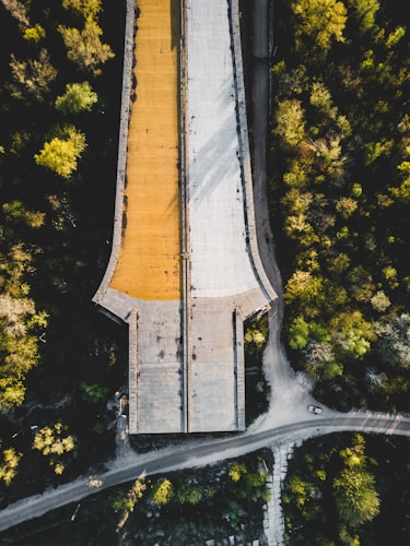 An aerial view of a partially constructed bridge surrounded by lush green trees. The bridge has two distinct sections with different surface colors; one is a light grey and the other is a warm orange. A road runs beneath the bridge, and a small vehicle is visible on the road.