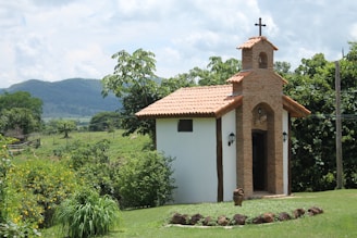 A small, rustic chapel with a tiled roof and a cross on top is set in the midst of lush greenery and rolling hills. The brickwork and simple architecture suggest a quaint, peaceful setting. Surrounding vegetation includes trees and bushes, with mountains visible in the distance against a partly cloudy sky.