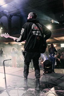 A musician is playing an electric guitar or bass on stage, facing away from the camera. The scene is dimly lit with blue and purple stage lights, creating a moody atmosphere. The audience in the background is partially visible, and stage equipment can be seen around the musician.