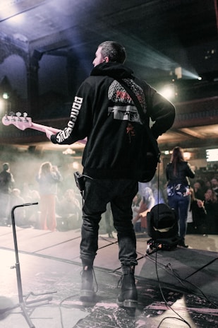 A musician is playing an electric guitar or bass on stage, facing away from the camera. The scene is dimly lit with blue and purple stage lights, creating a moody atmosphere. The audience in the background is partially visible, and stage equipment can be seen around the musician.