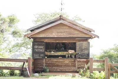 A small wooden roadside stall displays a variety of organic produce with chalkboard signs listing available items. The stall is surrounded by lush greenery, creating a rustic and inviting atmosphere. A wooden fence and a small plant in a pot are visible in front of the stall.