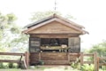 A small wooden roadside stall displays a variety of organic produce with chalkboard signs listing available items. The stall is surrounded by lush greenery, creating a rustic and inviting atmosphere. A wooden fence and a small plant in a pot are visible in front of the stall.