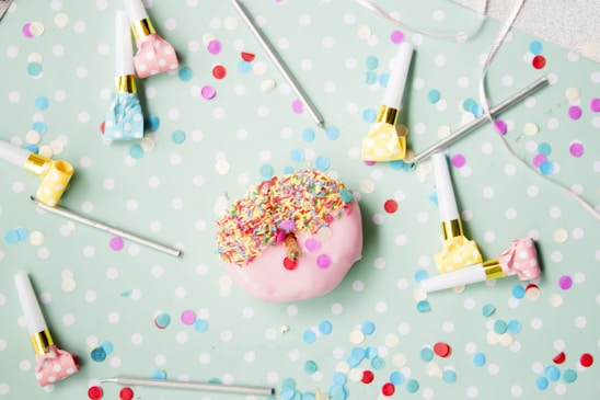 A pink donut covered in colorful sprinkles is centered on a pastel green polka dot surface. Surrounding the donut are several polka dot party blowers in pink, yellow, and blue. Scattered around are small, vibrant confetti pieces in various colors.